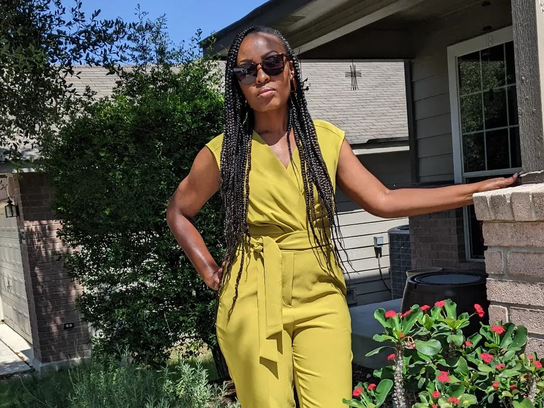 A woman in a yellow-green jumpsuit standing beside a porch and plants.